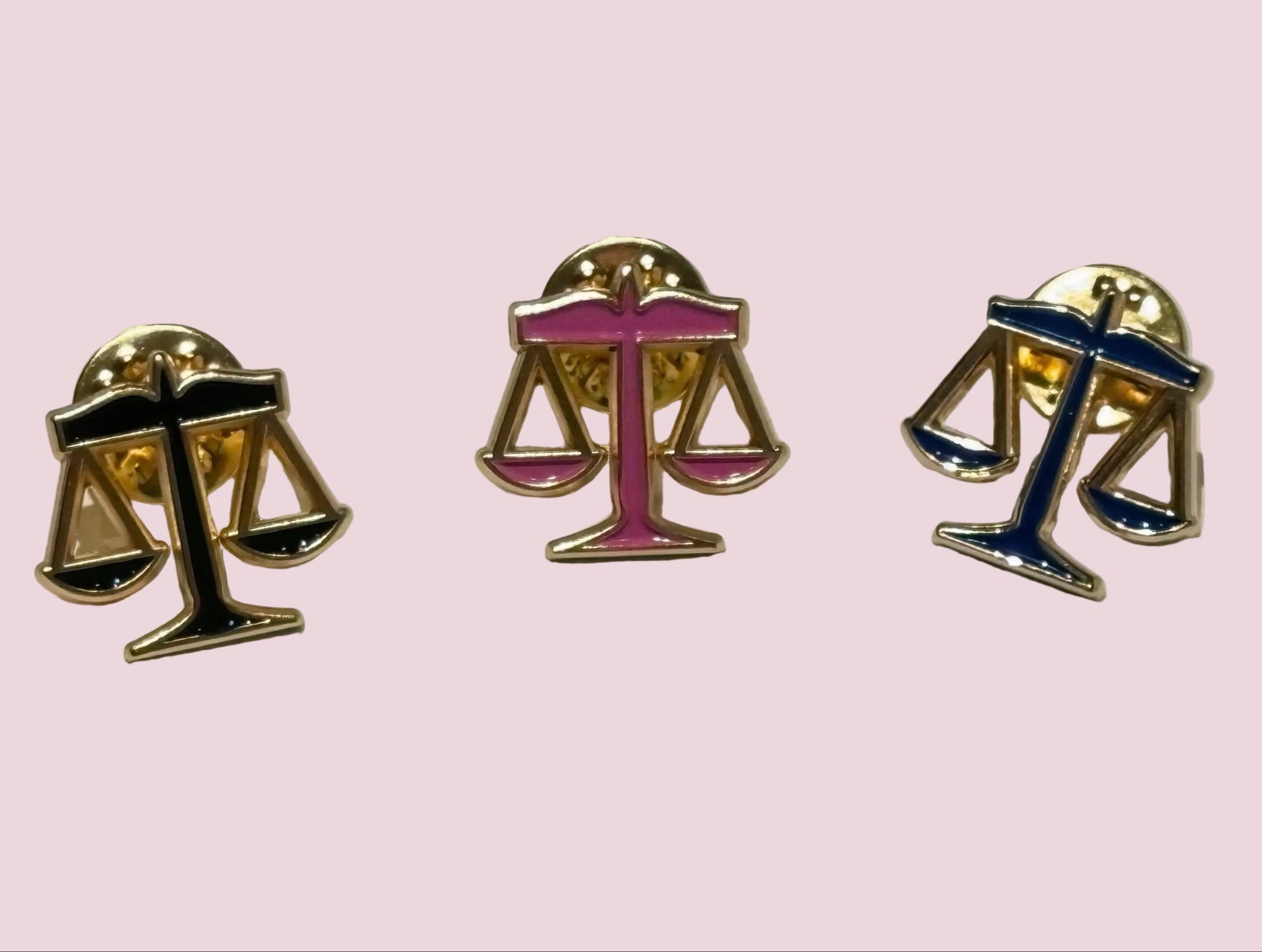 Three scales of justice lapel pins on a light pink background