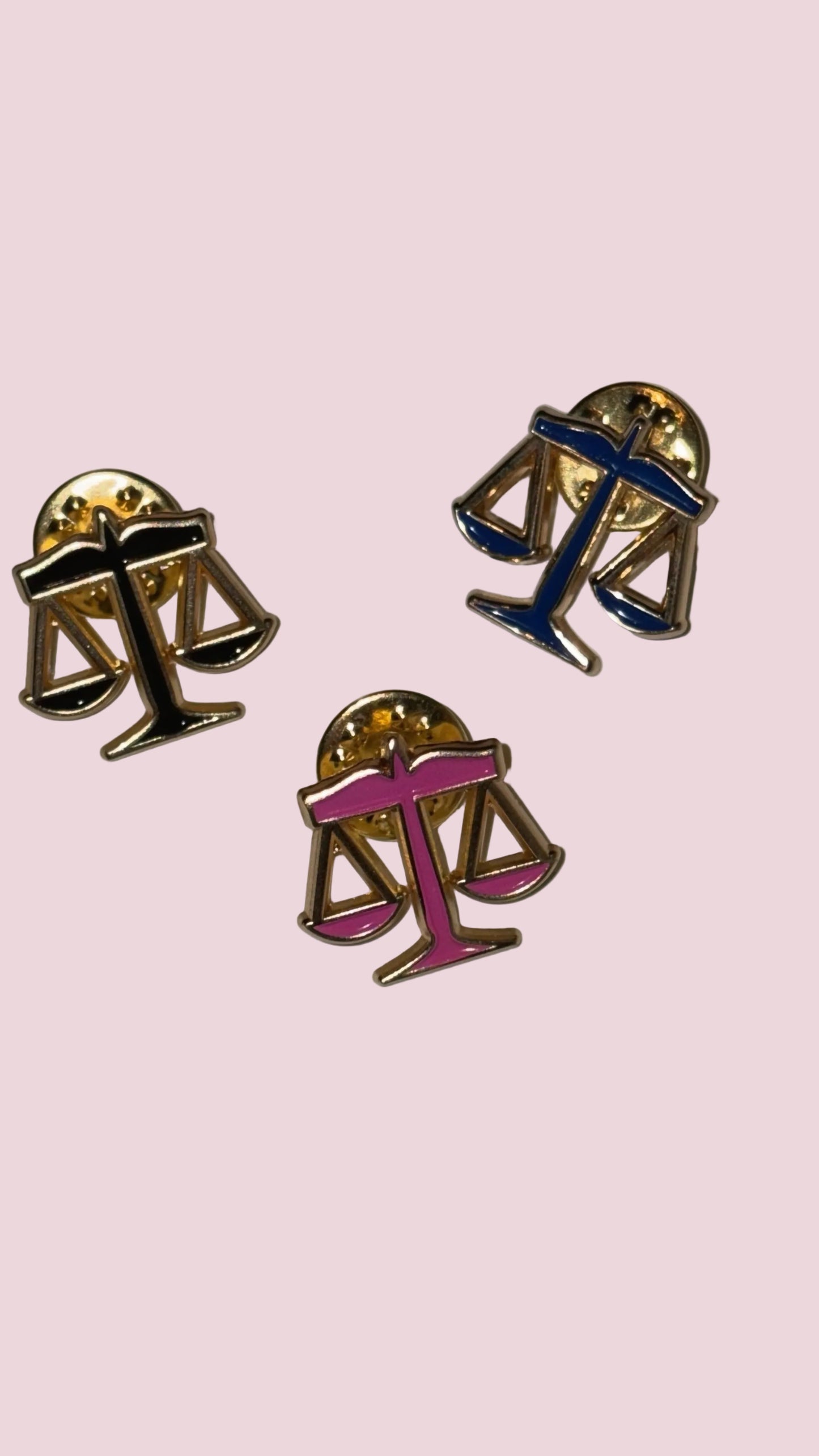 Three lapel pins with scales of justice on a light pink background.