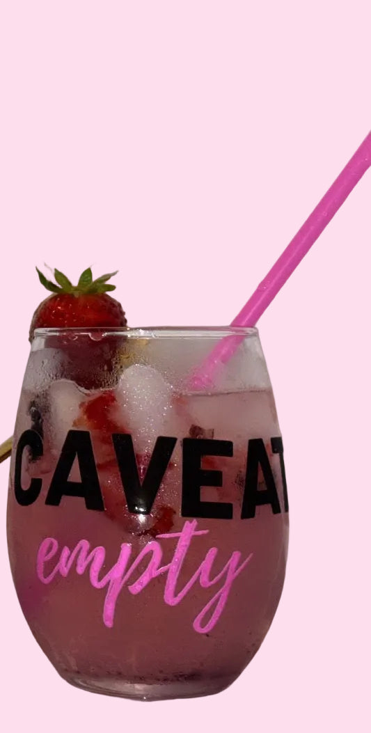 A clear stemless wine glass with black and pink text reading 'CAVEAT empty' sits next to a gray bowl of strawberries on a pink surface against a light background.