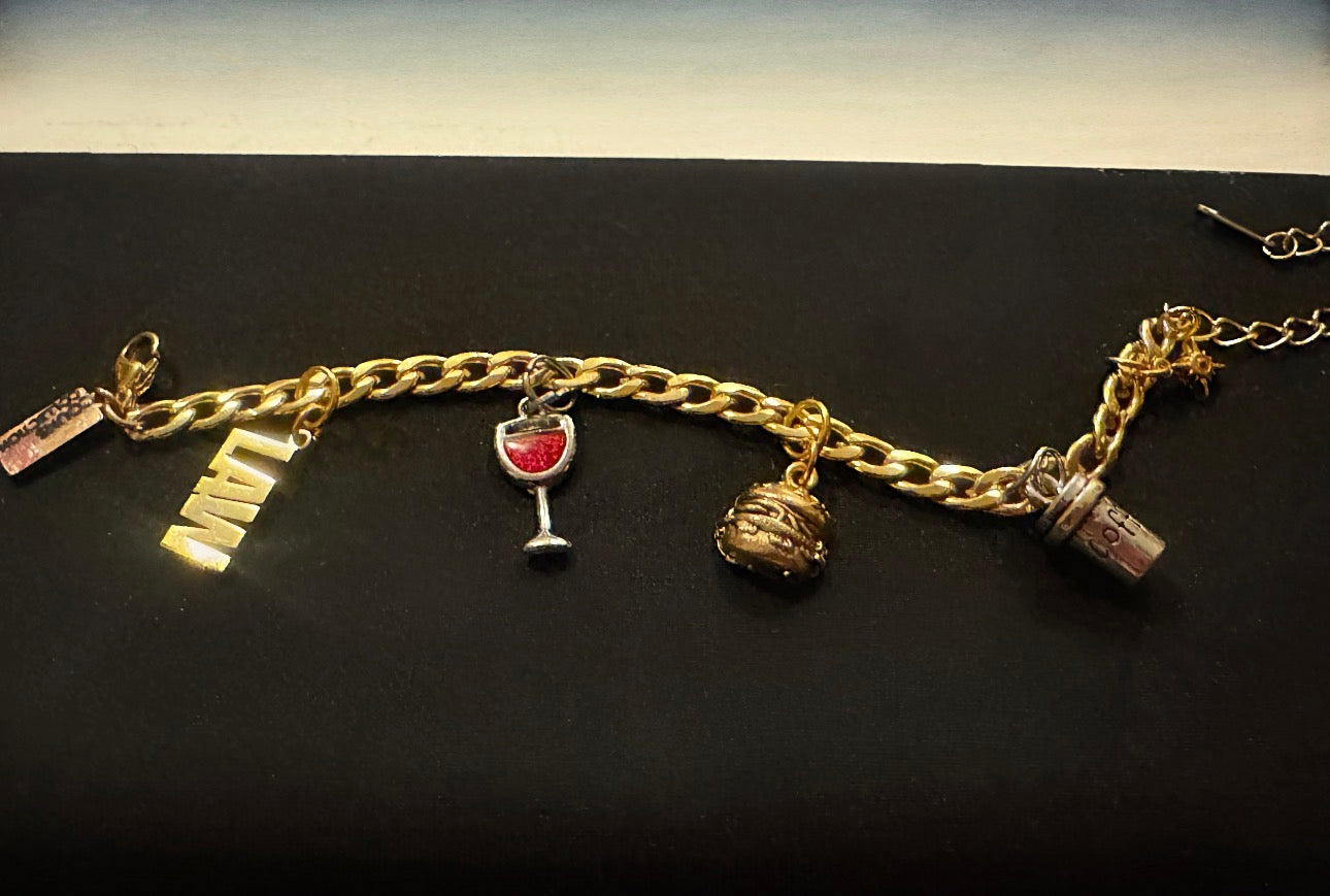 A gold-tone charm chain bracelet displayed horizontally on a dark background, featuring small charms including a the word “LAW”, a wine glass, a hamburger, a coffee cup, and a heart.