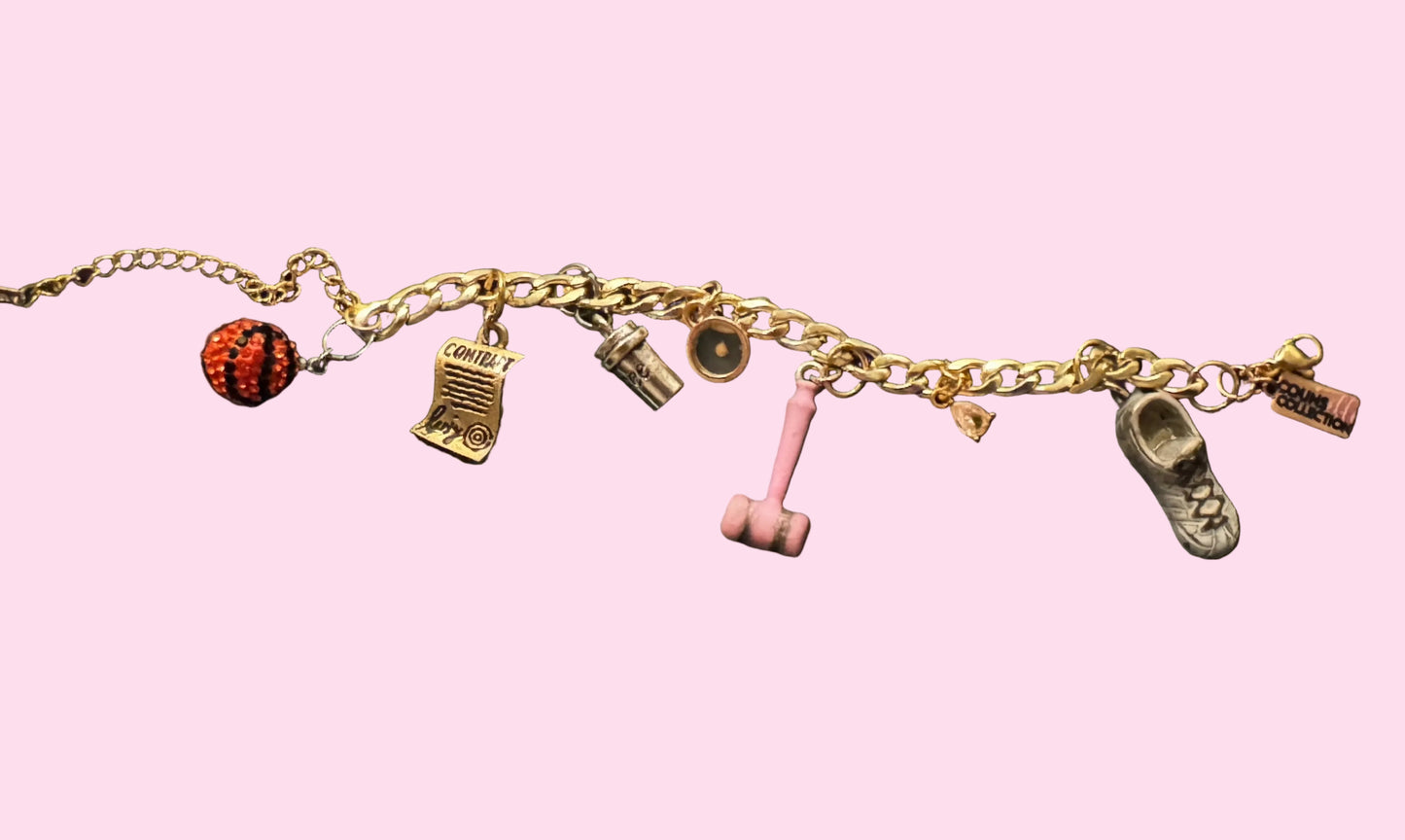 A gold-tone charm bracelet laid flat on surface, featuring multiple small charms including a black and orange bedazzled basketball, a signed contract charm, a mustard seed charm, a pink gavel charm, a white/bronze sneaker charm, and other small decorative charms.