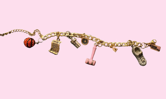A gold-tone charm bracelet laid flat on surface, featuring multiple small charms including a black and orange bedazzled basketball, a signed contract charm, a mustard seed charm, a pink gavel charm, a white/bronze sneaker charm, and other small decorative charms.