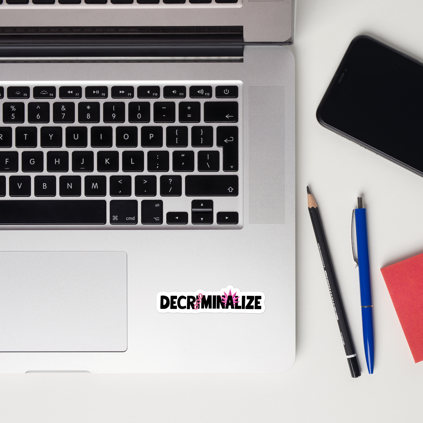 A sticker showing the text  "DECRIMINALIZE", with a broken pink chain over the first letter "I" and pink cannabis leaves behind the letter "A" ,  placed on a laptop. A pen, pencil, notepad, and mobile phone can also be seen on the flatlay. 