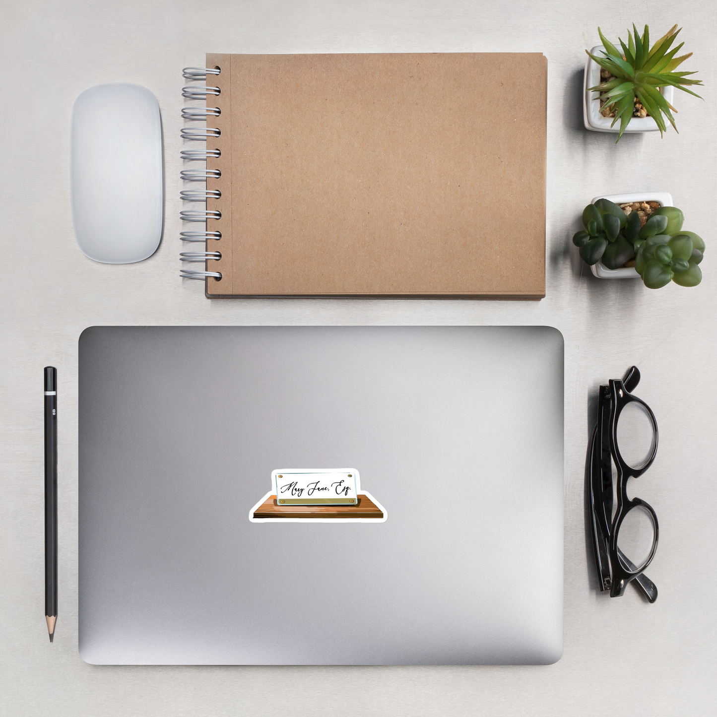 A sticker showing the text  "Mary Jane Esq" written on a nameplate placed on a laptop lid. A pencil, notebook, eyeglasses, computer mouse, and two potted succulents can also be seen on the flatlay.