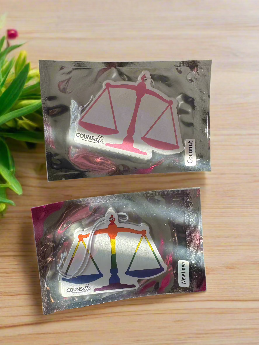 A picture of two car fresheners in silver packaging. The fresheners are in the shape of a justice scale and features the counselle collection® logo.