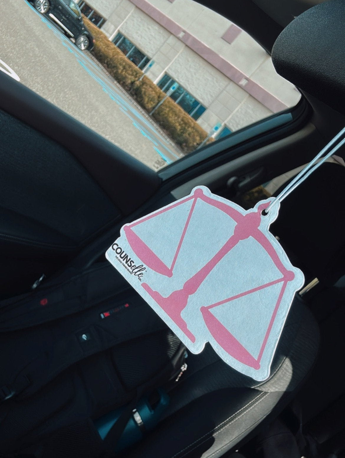 A picture of a car freshener hanging from a rearview mirror. The freshener is in the shape of a justice scale and features the counselle collection® logo.