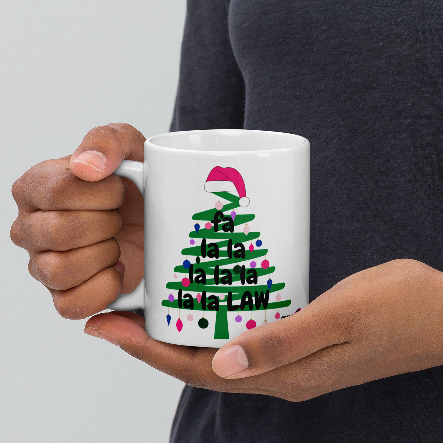 A person holding a white mug with an illustration of a green Christmas tree doodle with lights and a Santa hat on top, and the text "Fa La La La Law", with the counsellecollection logo at the bottom right. 