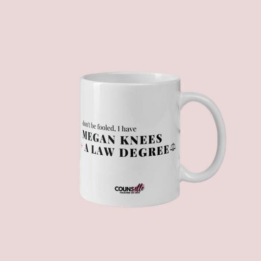 A ceramic mug sitting on a desk with a plant, a keyboard and two stacked books. The mug has the text "DON'T BE FOOLED, I'VE GOT MEGAN KNEES AND A LAW DEGREE" on the front. 