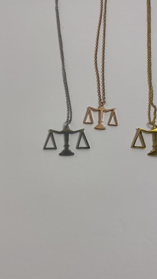 A stainless steel piece with 18k gold plating, featuring a sleek pendant in the shape of the scales of justice. Available in gold, rose gold, and silver.