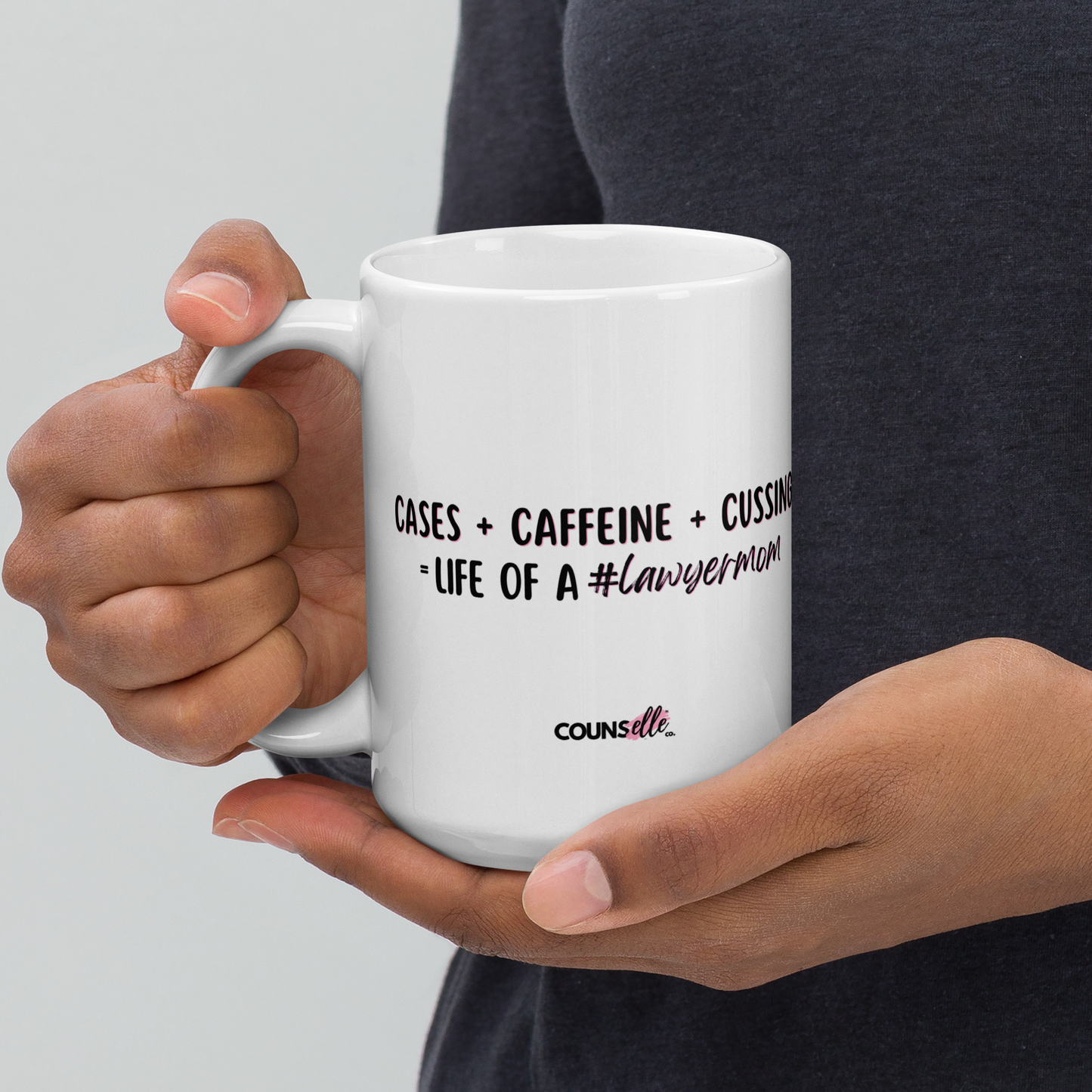 A person holding a 15oz white mug with the text "Cases + Caffeine + Cussing = LIFE OF A #lawyermom", and the counsellecollection logo at the bottom of the design