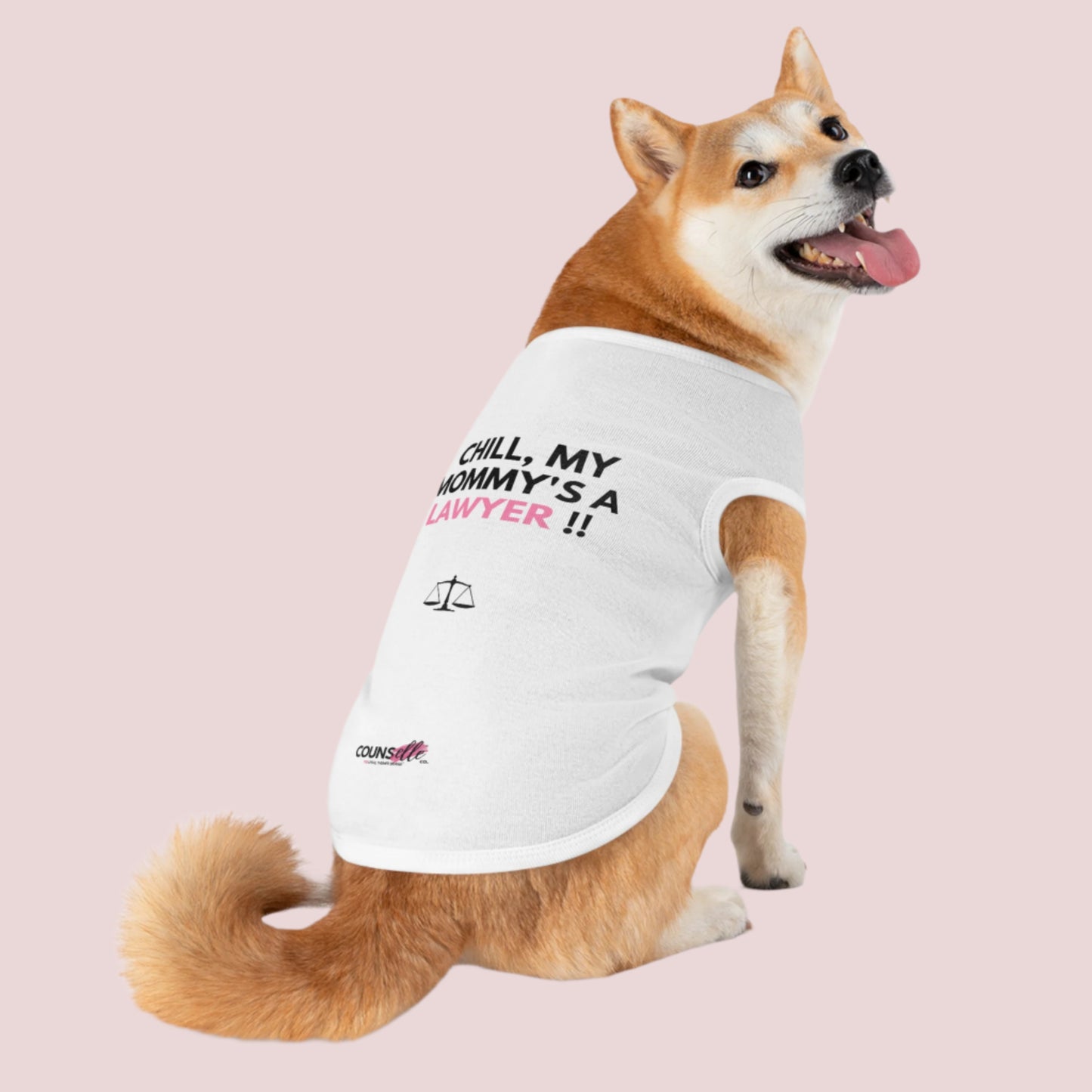 A dog wearing a white pet tee featuring the quote "Chill, my mommy's / daddy's a lawyer" in uppercase text, an illustration of justice scales below the text and the counsellecollection logo near the hem.