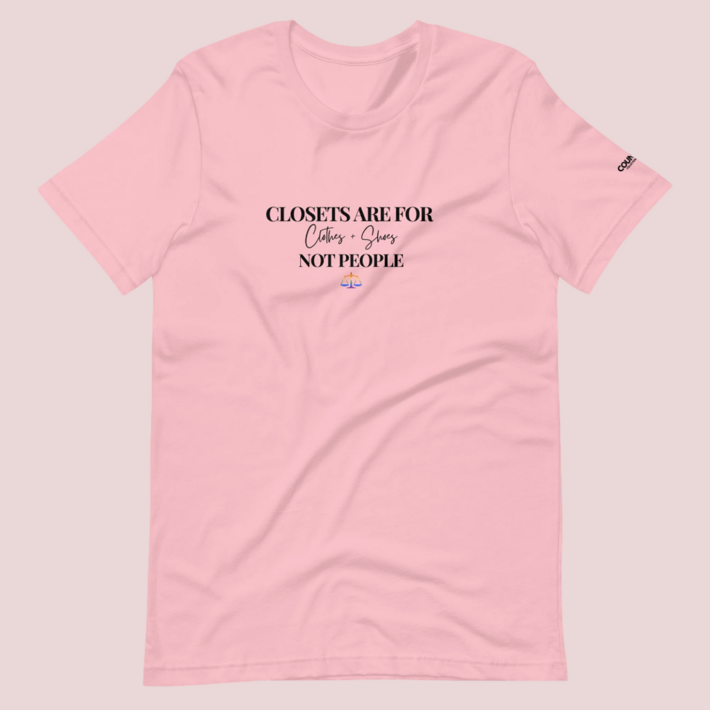 A pink crewneck tee with the text "CLOSETS ARE FOR CLOTHES + SHOES NOT PEOPLE" in black font, with an illustration of a rainbow justice scales underneath. The counsellecollection logo is seen on the left sleeve.