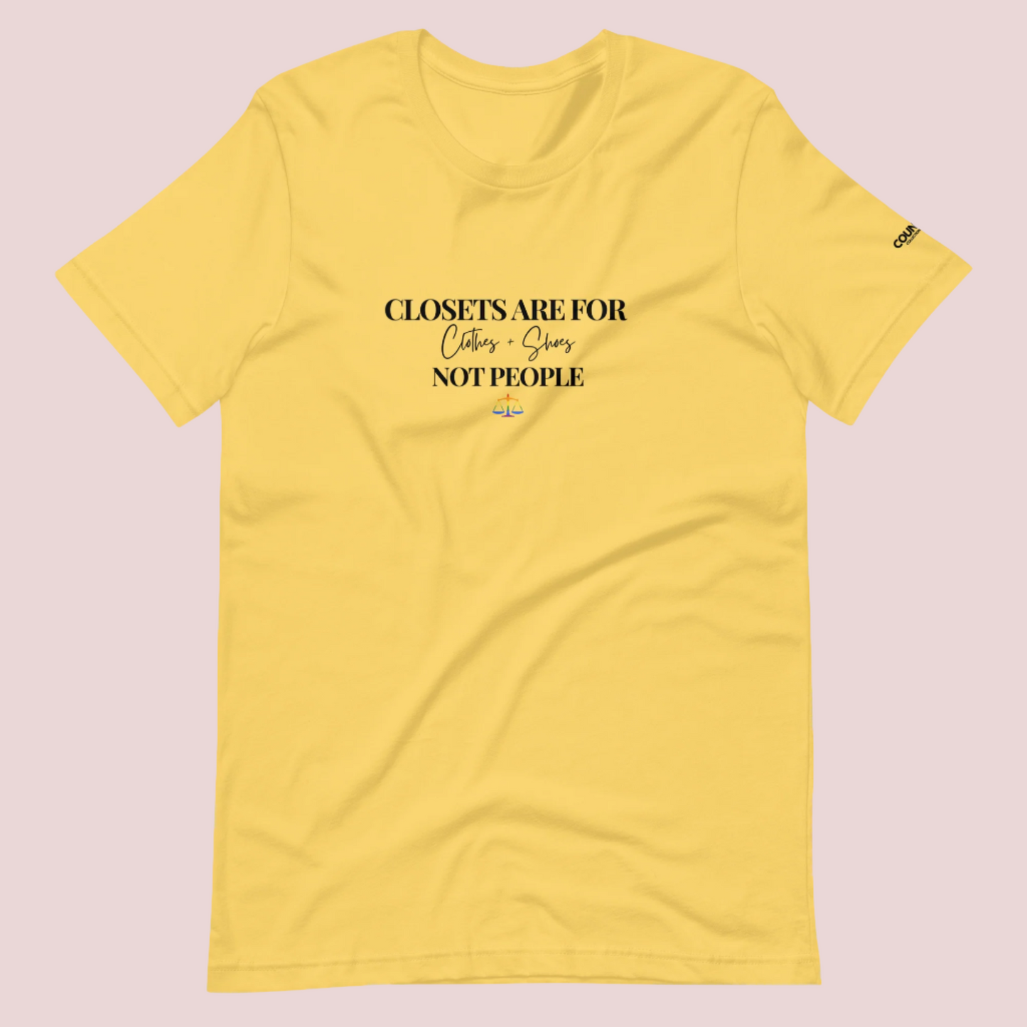 A yellow crewneck tee with the text "CLOSETS ARE FOR CLOTHES + SHOES NOT PEOPLE" in black font, with an illustration of a rainbow justice scales underneath. The counsellecollection logo is seen on the left sleeve.