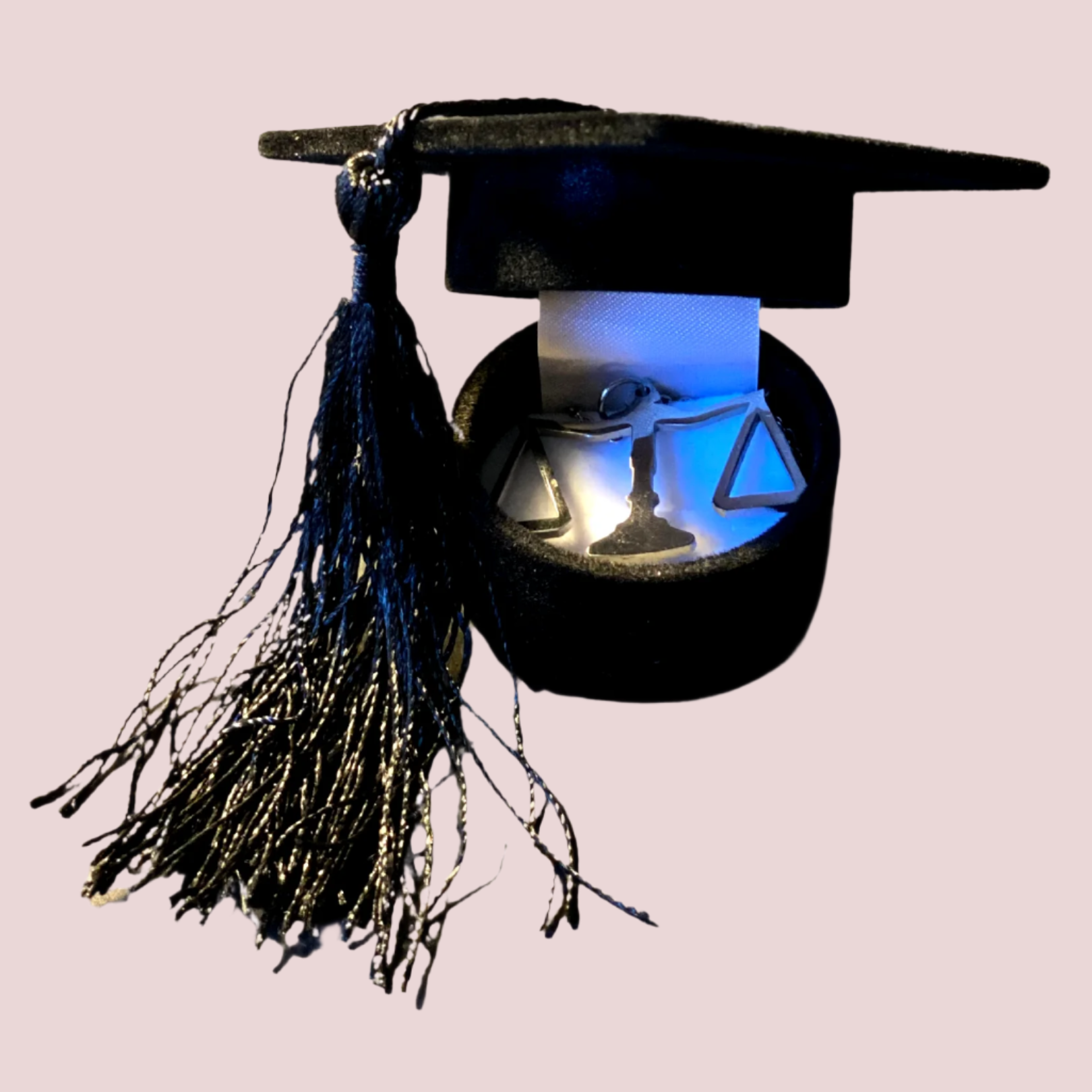  A fabric-lined, black jewelry box in the shape of a graduation cap with a black tassel on the side, showing a justice scales pendant inside