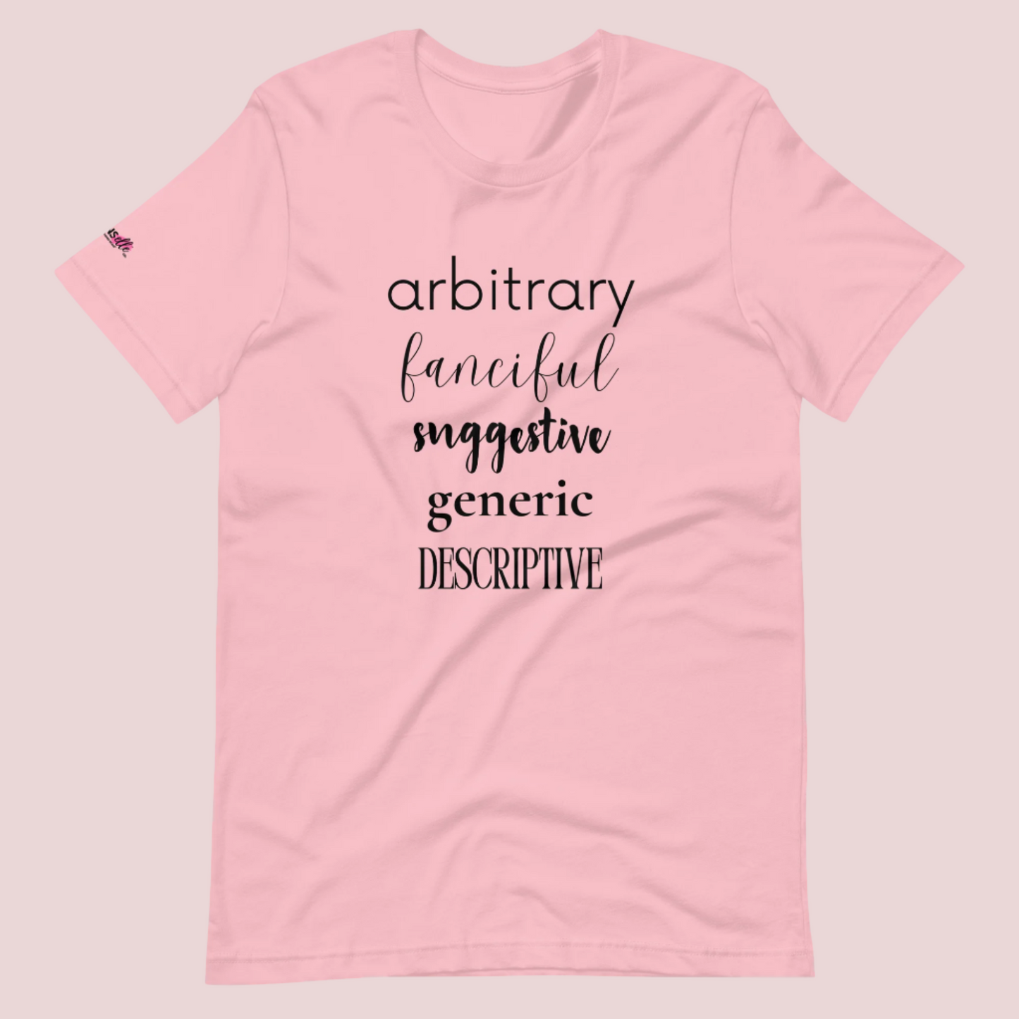 A pink roundneck tee featuring the words "arbitrary", "fanciful", "suggestive", "generic", and "descriptive" in different font styles, with the counsellecollection logo on a sleeve.