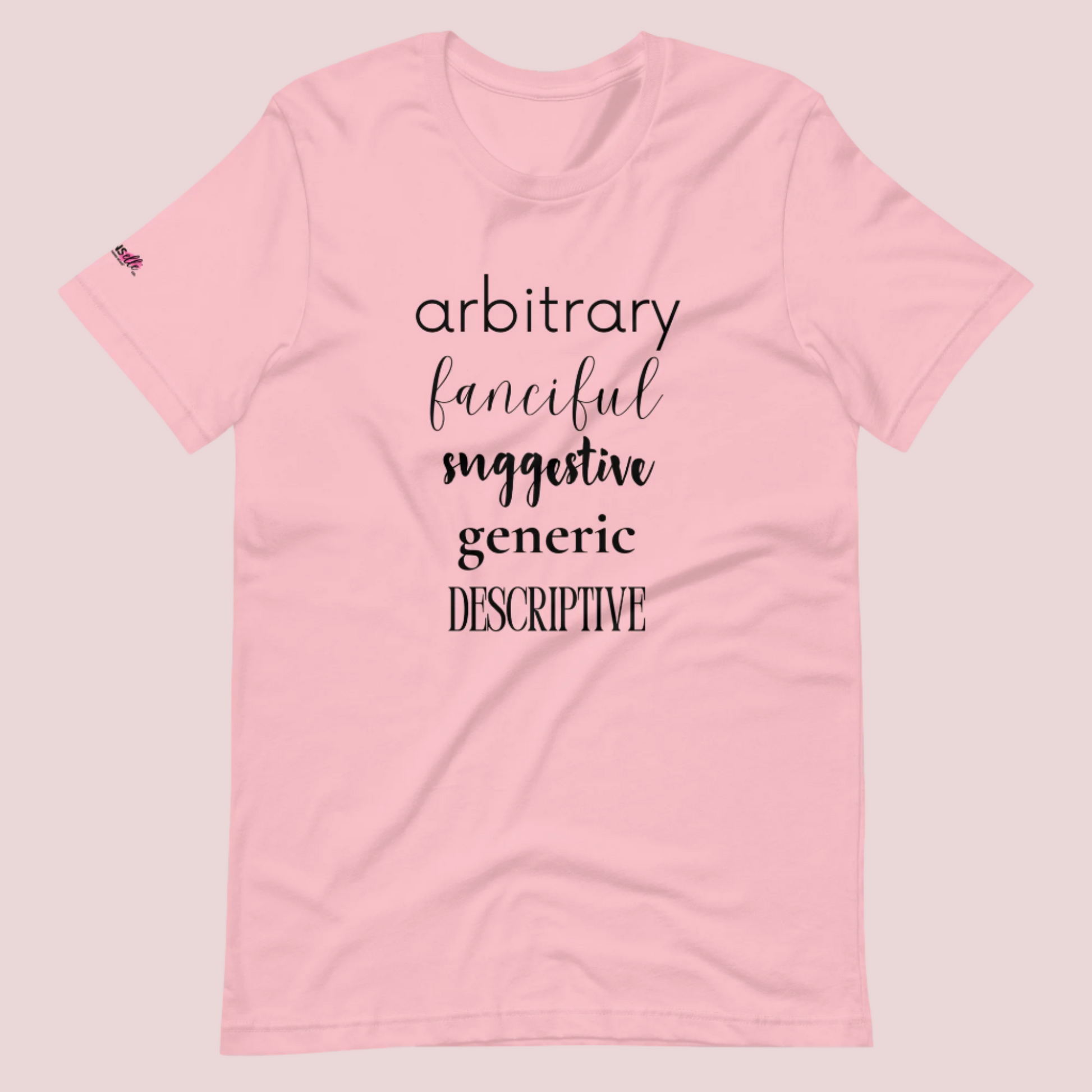 A pink roundneck tee featuring the words "arbitrary", "fanciful", "suggestive", "generic", and "descriptive" in different font styles, with the counsellecollection logo on a sleeve.