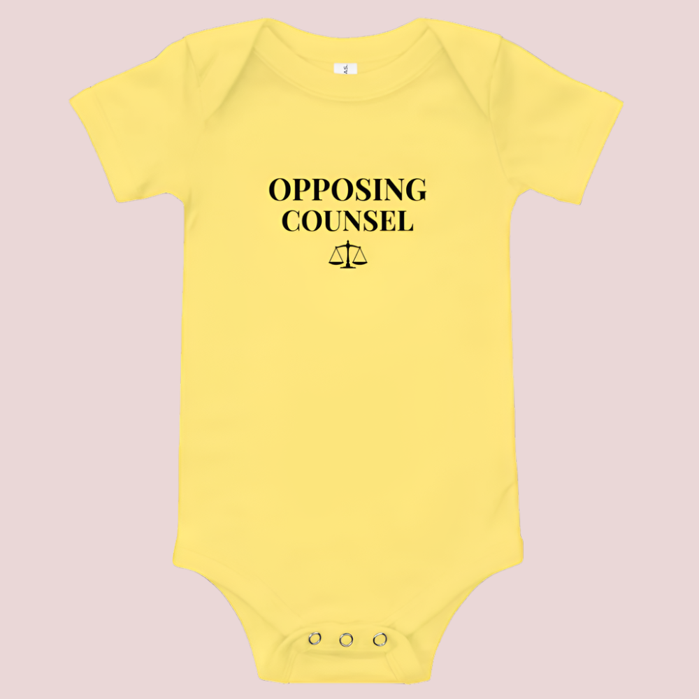 A yellow infant onesie featuring in black text, the words "OPPOSING COUNSEL" and an illustration of justice scales.
