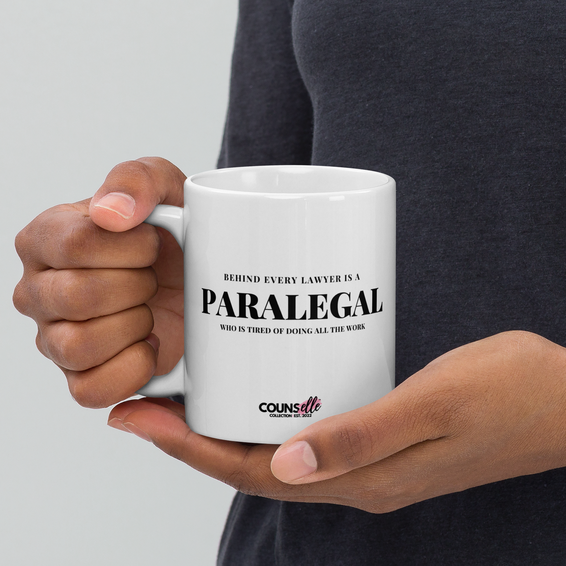 A person holding a white mug featuring the text "BEHIND EVERY LAWYER IS A PARALEGAL WHO IS TIRED OF DOING ALL THE WORK"