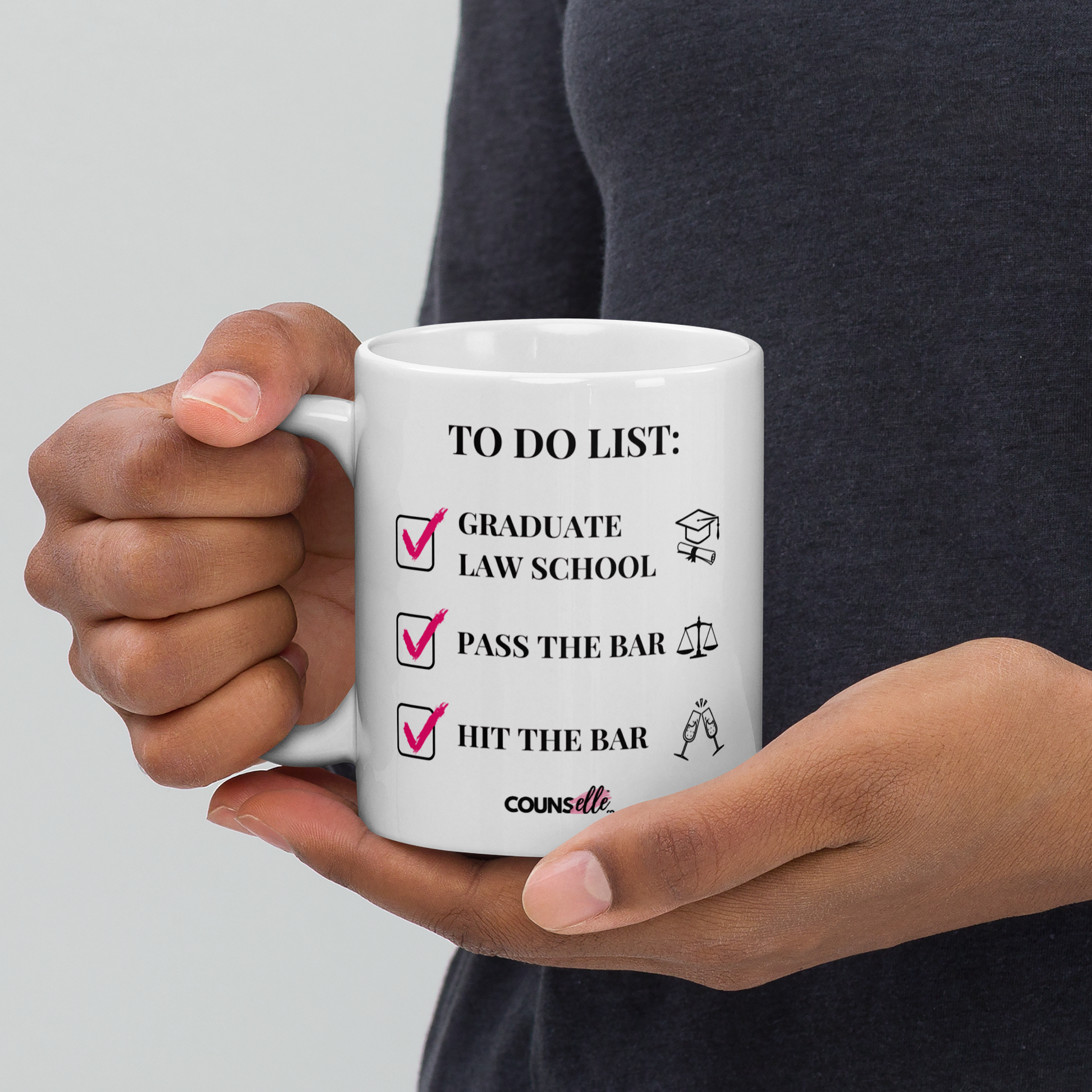  A person holding a white mug showing the text "TO DO LIST", with ticked-off boxes beside the texts "GRADUATE LAW SCHOOL" and an illustration of a graduation cap and diploma; "PASS THE BAR" ans an illustration of justice scales; and "HIT THE BAR" and an illustration of clinking champagne flutes. The counsellecollection logo is seen at the bottom of the design.